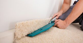 A person fitting a cream-colored carpet, using a carpet knife to carefully trim off excess material along the edge of the room.