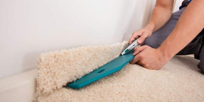 A person fitting a cream-colored carpet, using a carpet knife to carefully trim off excess material along the edge of the room.
